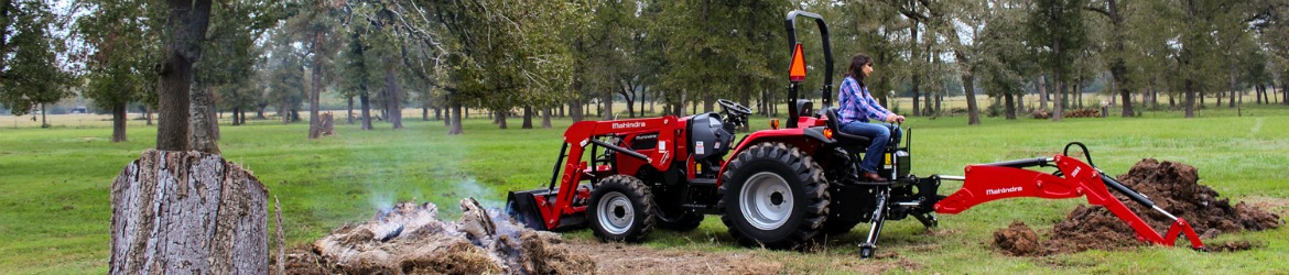 A Mahindra® Max Tractor digging in a field littered with coniferous trees.