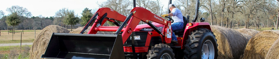 A man on a Mahindra® 4WD Tractor hauling large bails of hey on a field populated with trees.